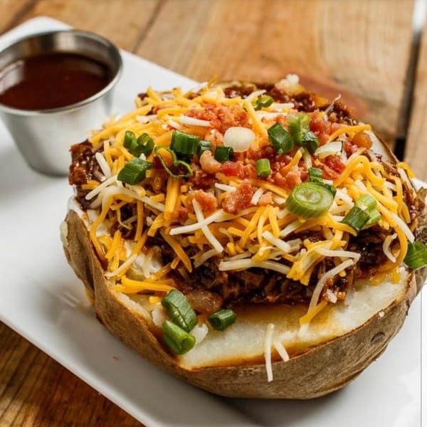 Loaded baked potato topped with shredded cheese, bacon bits, green onions, and a side of sauce.