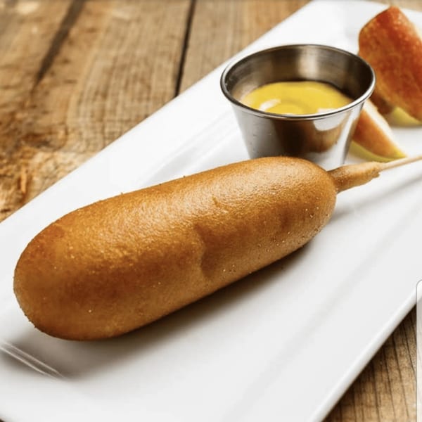 A golden-brown corn dog on a white plate with a side of mustard and apple slices.